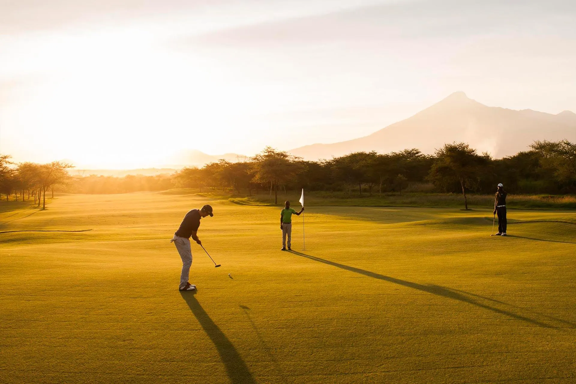 A golfer putting at sunrise, a caddie holding the flag, with a mountain beyond
