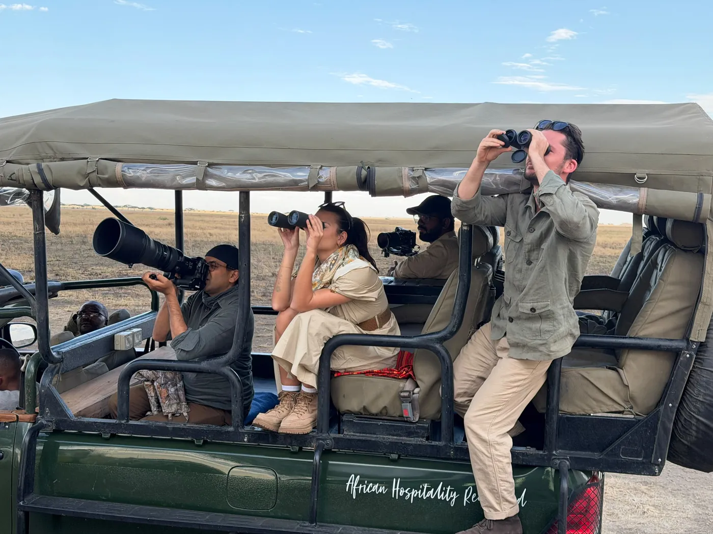 Guests with binoculars on an open safari vehicle, alongside photographers with long lenses